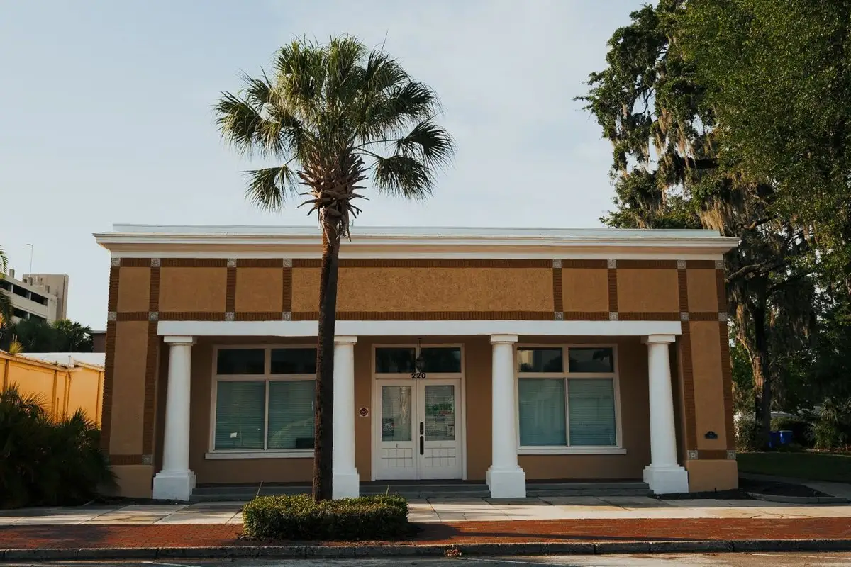 Tan stucco storefront with white columns, a sabal palm and a low hedge in front, and a live oak behind
