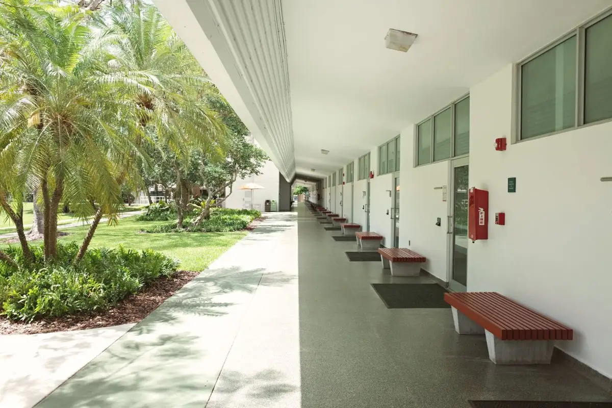Covered walkway along a low white building with garden beds, palms and benches